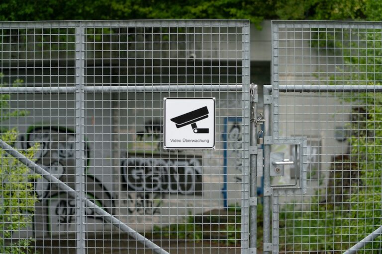 Verschlossenes Metallgittertor mit einem Schild, das eine Überwachungskamera zeigt und den Text "Video Überwachung" trägt; im Hintergrund eine mit Graffiti besprühte Wand und grüne Vegetation.