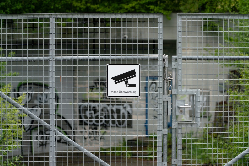 Verschlossenes Metallgittertor mit einem Schild, das eine Überwachungskamera zeigt und den Text "Video Überwachung" trägt; im Hintergrund eine mit Graffiti besprühte Wand und grüne Vegetation.