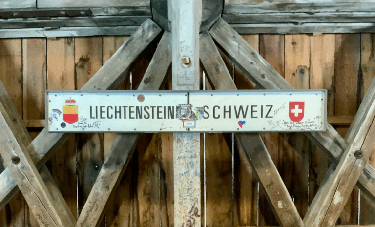 Grenztafel auf einer Holzbrücke mit der Aufschrift „Liechtenstein – Schweiz“, in der Mitte ein Punkt zur Markierung der Grenze. Links das Wappen von Liechtenstein, rechts das Schweizerkreuz. Die Tafel ist von zahlreichen handschriftlichen Notizen und Kritzeleien umgeben, die Holzkonstruktion der Brücke ist deutlich sichtbar.