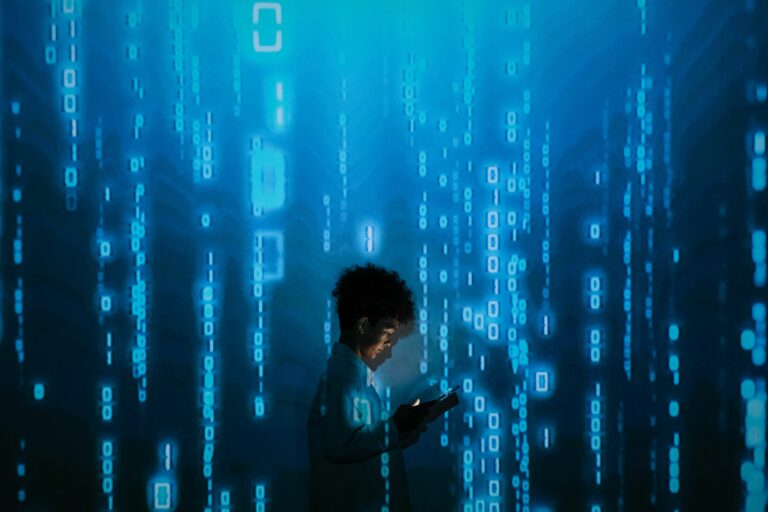 Das Bild zeigt eine dunkelhaarige Person im Profil, die in einem hellen Hemd vor einer leuchtenden blauen Wand steht. Die Person hält ein kleines elektronisches Gerät oder Smartphone in den Händen, das ihr Gesicht und ihre Hände sanft beleuchtet. Im Hintergrund ist ein digitaler Matrix-Effekt zu sehen: vertikale Spalten aus blauen Einsen, Nullen und Code-Zeichen, die den Raum in ein kühles, technologisches Licht tauchen und eine futuristische Atmosphäre von Datenverarbeitung und Programmierung erzeugen.