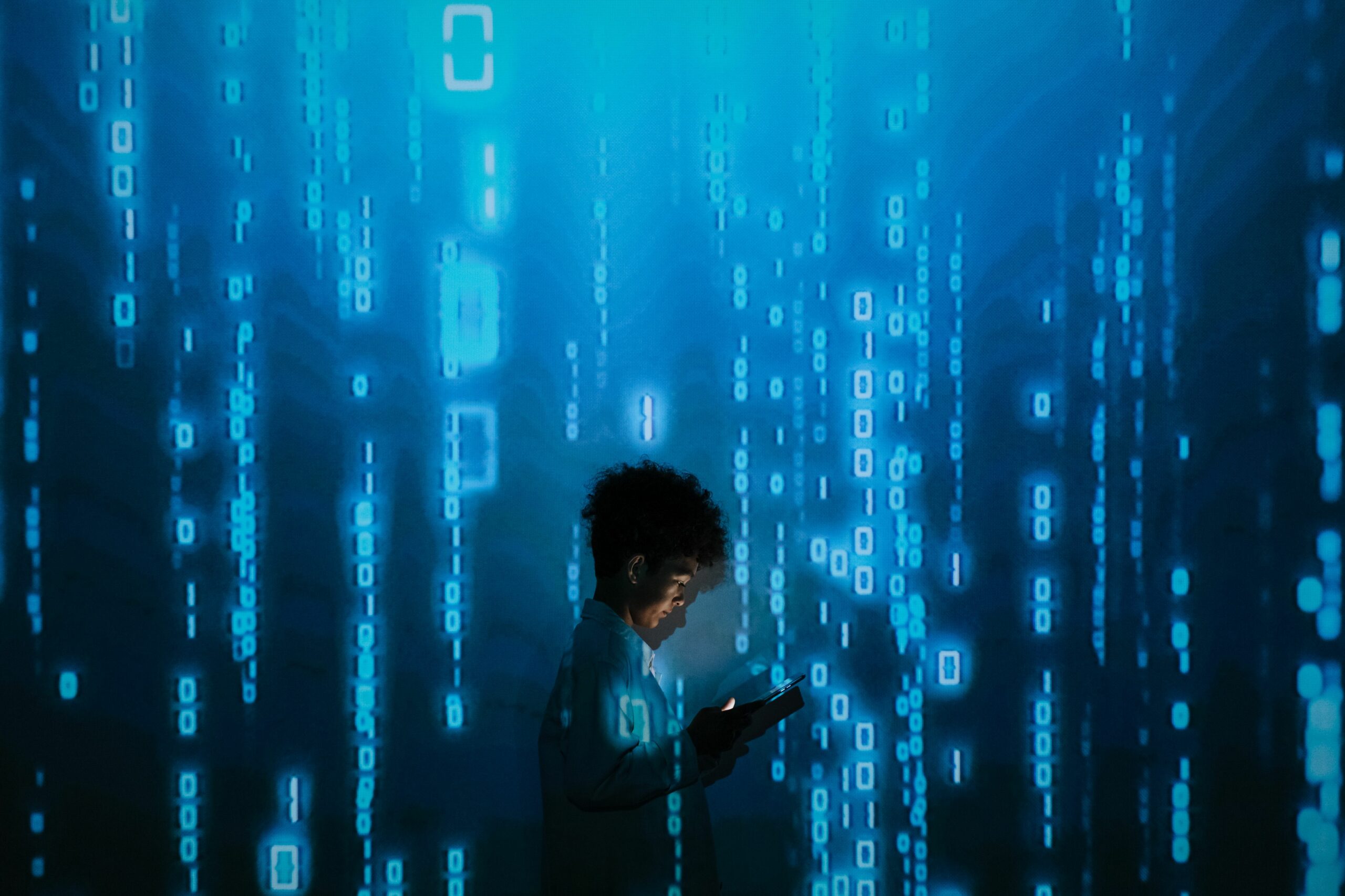 Das Bild zeigt eine dunkelhaarige Person im Profil, die in einem hellen Hemd vor einer leuchtenden blauen Wand steht. Die Person hält ein kleines elektronisches Gerät oder Smartphone in den Händen, das ihr Gesicht und ihre Hände sanft beleuchtet. Im Hintergrund ist ein digitaler Matrix-Effekt zu sehen: vertikale Spalten aus blauen Einsen, Nullen und Code-Zeichen, die den Raum in ein kühles, technologisches Licht tauchen und eine futuristische Atmosphäre von Datenverarbeitung und Programmierung erzeugen.