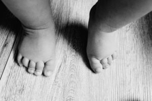 Das in Schwarz-Weiß gehaltene Bild zeigt die kleinen, pausbäckigen Füsse eines Kleinkindes oder Babys von oben. Die Füsse stehen auf einem hellen Holzboden mit sichtbarer Maserung. Die Perspektive betont die kleinen Zehen und die typischen Speckröllchen an den Knöcheln.