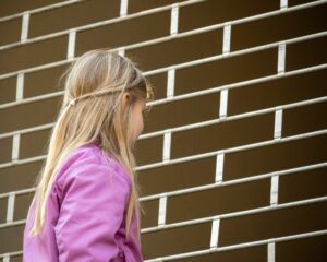 Das Bild zeigt ein junges Mädchen mit langem, blondem Haar in einer Seitenansicht von hinten, vor einem Fallgitter stehend. Sie trägt eine auffällige pinke oder violette Jacke. Ihr Haar ist am Hinterkopf teilweise zusammeng