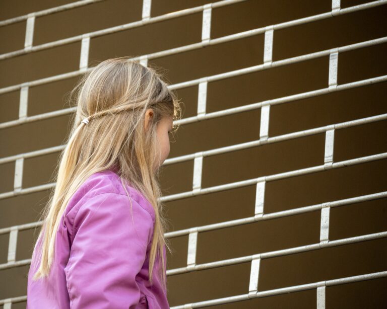 Das Bild zeigt ein junges Mädchen mit langem, blondem Haar in einer Seitenansicht von hinten, vor einem Fallgitter stehend. Sie trägt eine auffällige pinke oder violette Jacke. Ihr Haar ist am Hinterkopf teilweise zusammeng