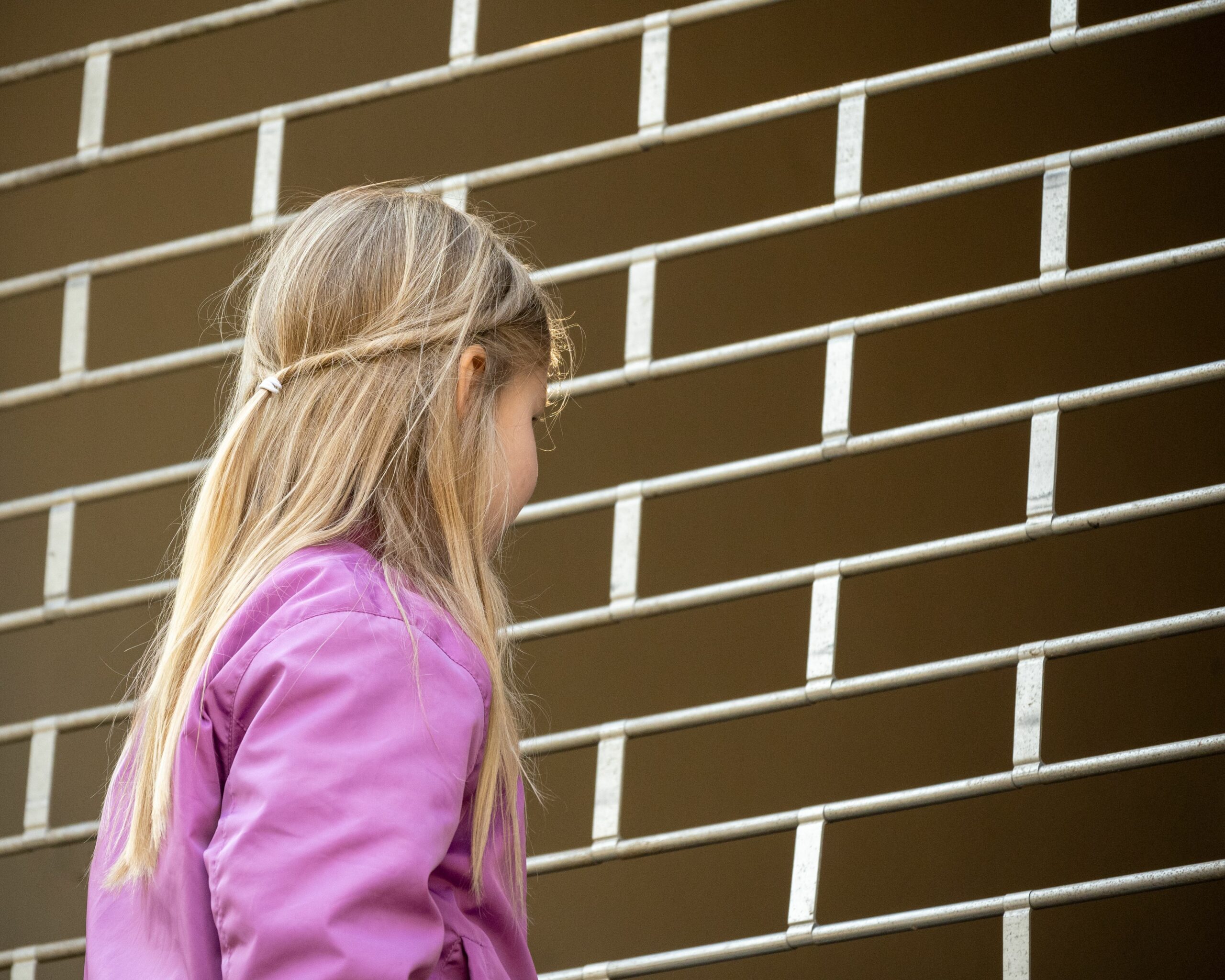 Das Bild zeigt ein junges Mädchen mit langem, blondem Haar in einer Seitenansicht von hinten, vor einem Fallgitter stehend. Sie trägt eine auffällige pinke oder violette Jacke. Ihr Haar ist am Hinterkopf teilweise zusammeng
